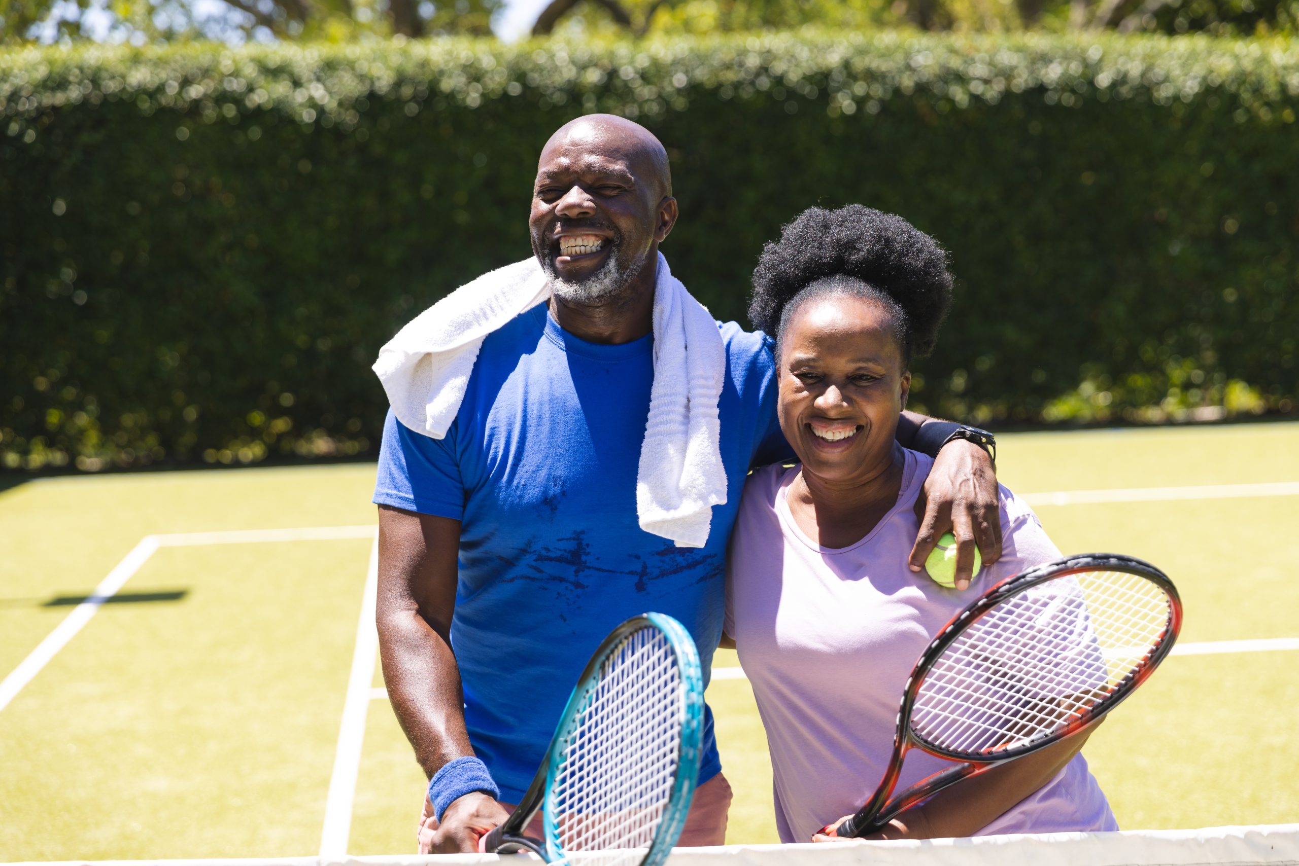 Active and happy mature couple holding tennis rackets on an outdoor tennis court.